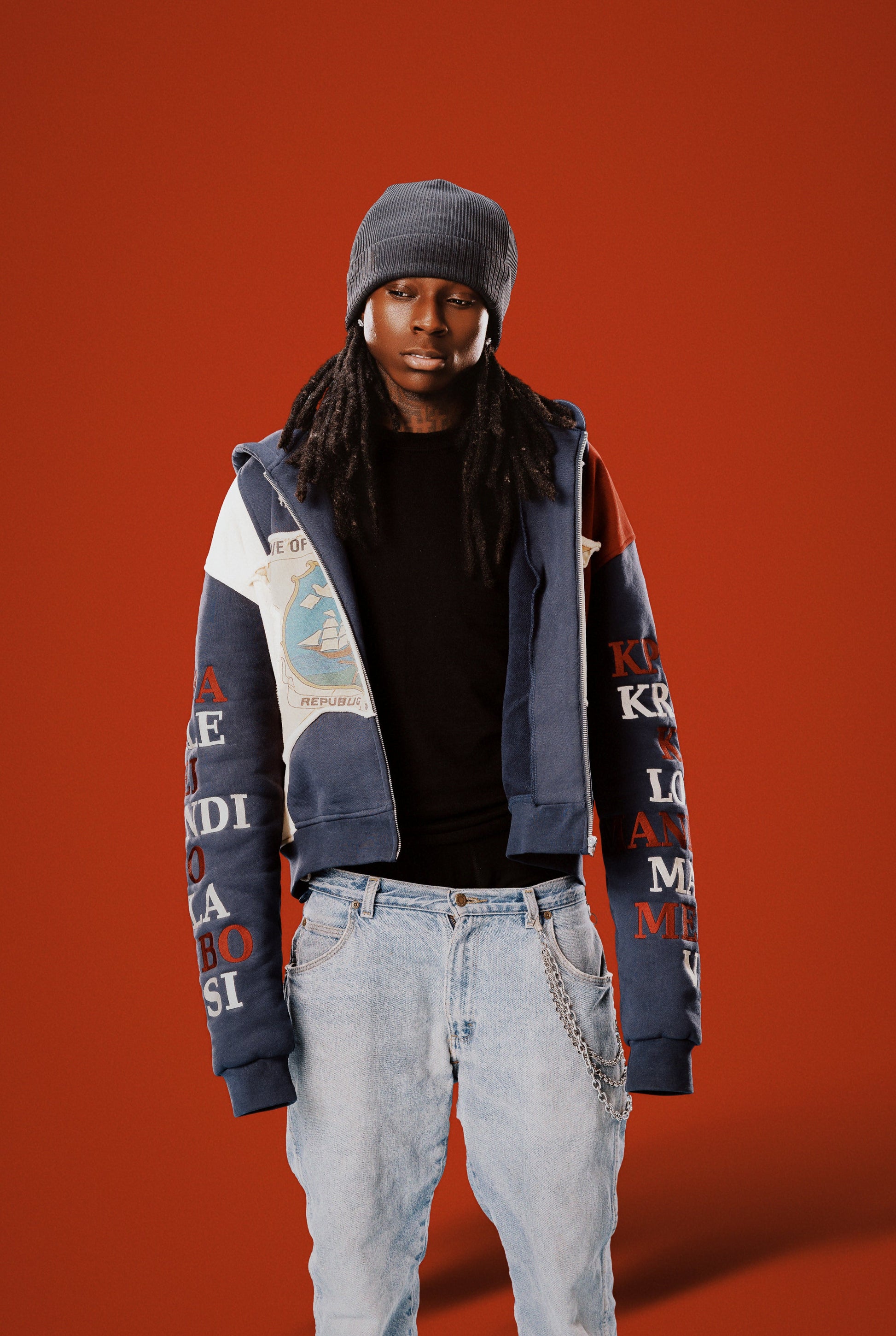 Person wearing a colorful jacket with text, black beanie, and light jeans on an orange background