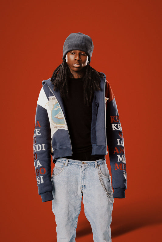 Person wearing a colorful jacket with text, black beanie, and light jeans on an orange background